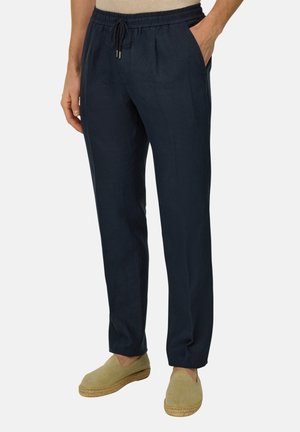 Hombre vestido con pantalones casuales azul marino con cordón y bolsillos, y alpargatas beige sin cordones, de pie frente a un fondo liso.