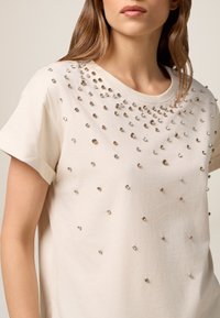 Tricou din bumbac bej cu mâneci scurte, având aplicate ace de metal aurii și argintii, creând o suprafață texturată, decorativă.