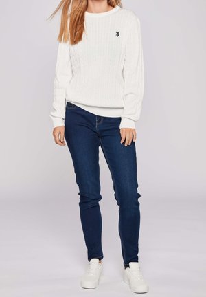 Valkoinen palmikkoneulepusero pyöreällä kaula-aukkolla ja resorihihoilla, yhdistetty tummansinisiin slim fit -farkkuihin ja valkoisiin tennareihin.