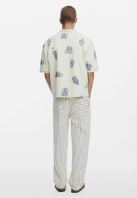 Rövid ujjú fehér ing kék és sárga absztrakt mintákkal, amit krémcsíkos, bő szárú nadrággal párosítanak, hátul zsebekkel.