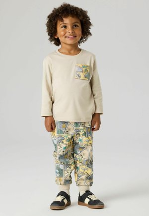 Ungt barn med krøllet hår iført beige langærmet skjorte og farverige bukser med dinosauermønster, smilende foran en ensfarvet grå baggrund.