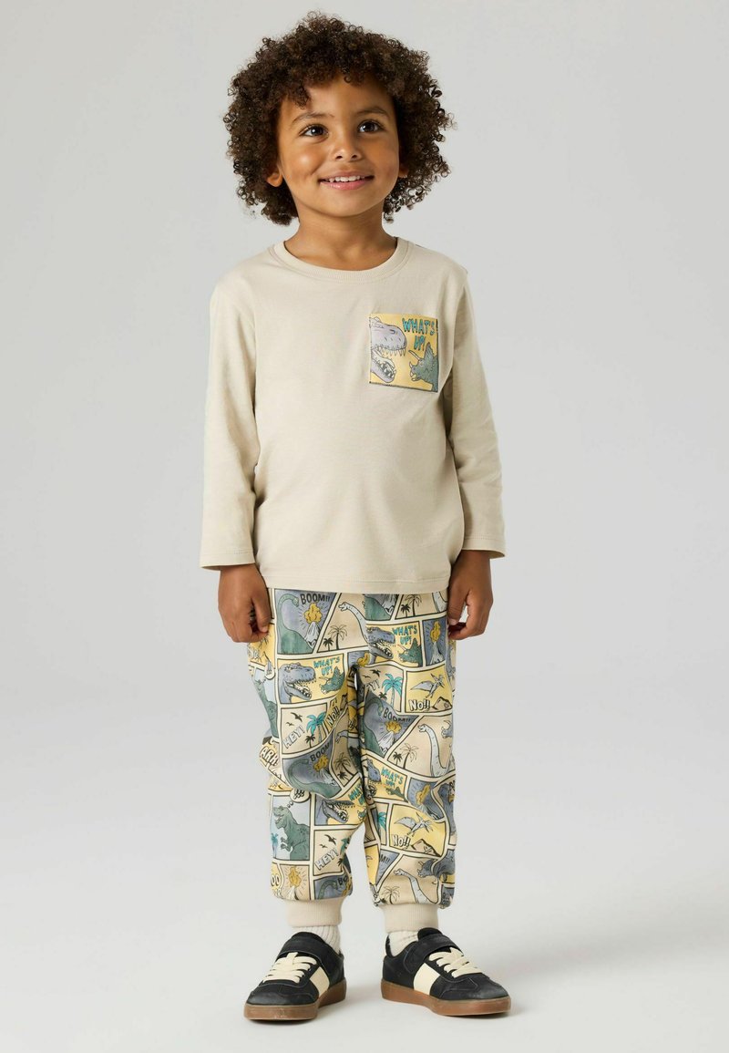 Małe dziecko z kręconymi włosami, ubrane w beżową bluzkę z długim rękawem i kolorowe spodnie z wzorem dinozaurów, uśmiecha się na tle jednolitego szarego tła.