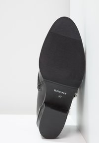 Botín de cuero negro con suela de goma texturizada, punta redonda, cremallera trasera y marca grabada "BRONX 37" en la parte inferior.