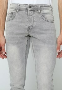 Jeans en denim gris avec une coupe slim et une texture délavée. Comprend cinq poches et une fermeture à bouton sur le devant. Couture de couleur contrastée.
