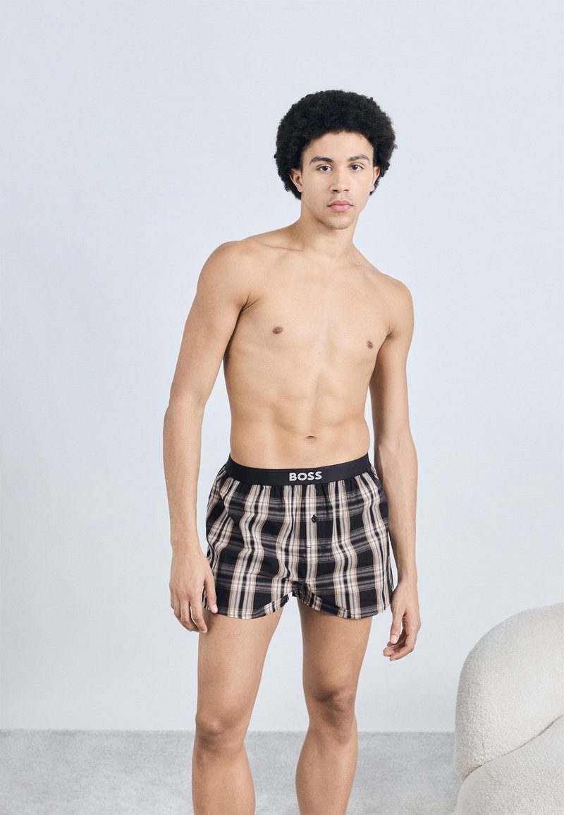 Fekete és bézs kockás boxeralsó elasztikus derékpánttal, amelyen a "BOSS" logó látható, egy álló modell viseli.