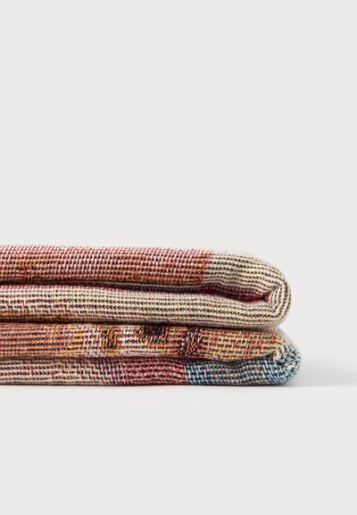 Deux pièces textiles tissées et pliées avec des fils rouges, blancs, jaunes, noirs et bleus empilées sur une surface blanche.