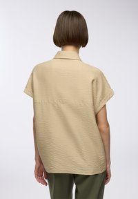 Camicia beige, con texture, colletto, maniche corte e vestibilità rilassata. La vista posteriore mostra cuciture sottili e un orlo posteriore leggermente più lungo.