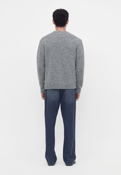 Homme debout de dos, portant un pull gris tricoté et un jean bleu ample sur un fond clair uni.