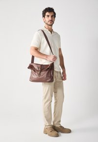 Brown leather messenger bag with a flap and a metal clasp, worn crossbody over a cream textured polo shirt and beige pants.