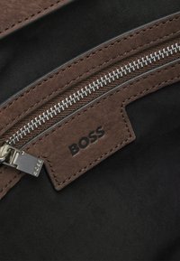 Bruine leren ritsluiting met zilverkleurige hardware, met een zwarte stofachtergrond en een gestikerde label met de opdruk "BOSS."