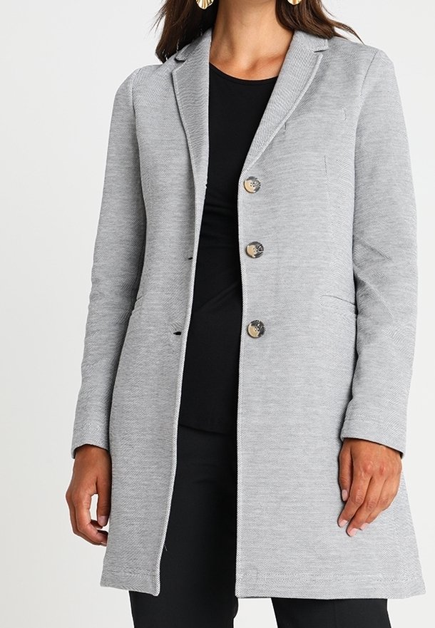 Manteau long gris en tissu texturé, avec un col à revers cranté, trois boutons et des poches latérales. Porté sur un haut noir.