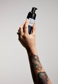 Bottiglia in bianco e nero etichettata "Mousse per Volume al Sale Marino", tenuta in una mano con intricati tatuaggi, dotata di dosatore a pompa.