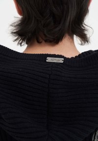 Maglione in maglia nera a coste con un'etichetta argentata sul retro del colletto. Il tessuto ha una consistenza morbida con righe orizzontali visibili.