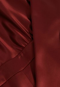 Burgundijska satenska tkanina s glatkom teksturom, drapiranim detaljima i naglaskom na šavu. Prikaz blizu preklopa tkanine.
