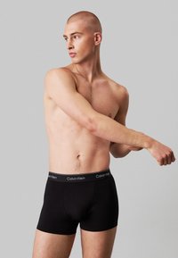 Zwarte boxershorts met een merken elastische tailleband. Het model heeft een geschoren hoofd en poseert, waardoor de pasvorm en textuur van de stof worden getoond.