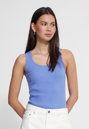 Jeune femme aux longs cheveux foncés portant des boucles d'oreilles en perles, un haut bleu clair sans manches et un pantalon blanc, debout les mains derrière le dos.