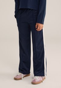 Cilvēks, kurš uzvilcis tumši zilo sporta bikses ar baltām sānu svītrām un rozā zamšādas sneakerus ar baltām šņorēm, stāv uz gaiša fona.