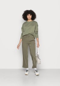 Sweatshirt verde-oliva com um corte descontraído, combinado com calças largas a condizer. Ténis brancos e uma bolsa cinza-clara completam o look.