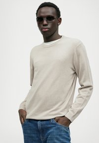 Maglione beige a girocollo in maglia testurizzata, abbinato a jeans blu in denim. Modella con occhiali da sole scuri in piedi con le mani in tasca.