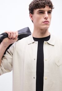 Νεαρός άνδρας που φοράει μια μπεζ πλεκτή μπλούζα πάνω από μαύρο τοπ, κουβαλώντας μια μαύρη τσάντα στον ώμο, κοιτάζει στο πλάι με ουδέτερη έκφραση.