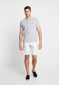Svetlošedá polo košeľa s golierom, krátkymi rukávmi a zeleným logom, v kombinácii s bielymi šortkami a námorníckymi slip-on topánkami.