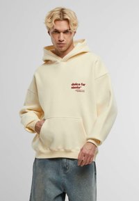 Hoodie giallo chiaro con tasca frontale. Presenta un logo stampato in rosso scuro sul lato sinistro del petto. Vestibilità ampia e tessuto di peso medio.