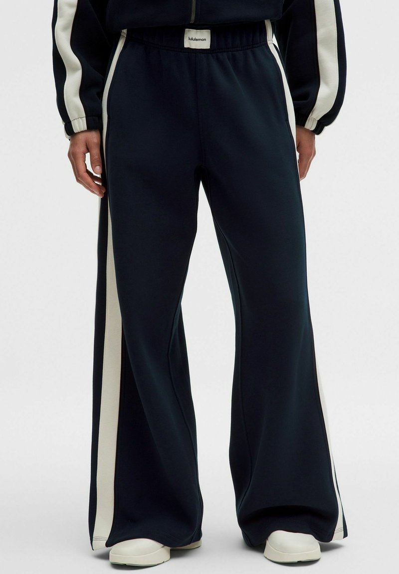 Tmavě modré široké kalhoty s bílými bočními pruhy, elastickým pasem a bílými teniskami, nošené osobou s uvolněnými rukama po stranách.