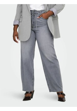 Nő, magasderekú, szürke, bőszárú farmert, világosszürke csíkos blézert visel, az ujjait felhajtva, fehér felsővel és fekete magassarkú cipővel.