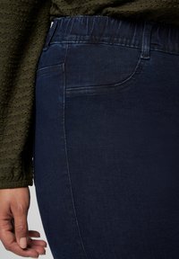 Jean en denim bleu foncé avec une texture lisse, taille élastique et poches latérales. Gros plan de la zone des hanches, mettant en avant les détails de couture.