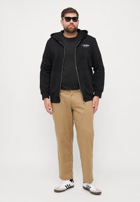 Zwarte hoodie met ritssluiting en voorzakken, gedragen over een zwart t-shirt. Beige broek en witte schoenen met zwarte strepen. Model draagt een zonnebril.
