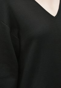 Maglione nero a V con una texture liscia e morbida, caratterizzato da una vestibilità morbida e spalle leggermente scese. Design classico e minimalista.