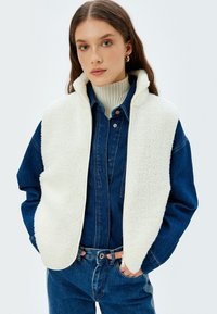 Ein weißer Fleece-Westen über einem Jeanshemd, mit hohem Kragen, Reißverschluss und Seitentaschen. Die Weste hat eine weiche Textur und keine Muster.
