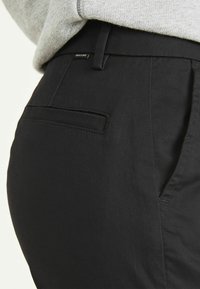 Pantaloni neri realizzati in un tessuto liscio, dotati di una tasca laterale e una vita piatta con un piccolo etichetta del marchio. Vestibilità sartoriale con cuciture ordinate.