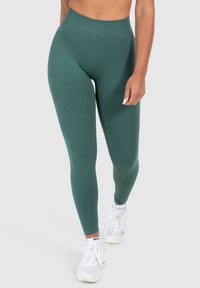 Leggings verdi a vita alta in un materiale liscio e elasticizzato, caratterizzati da un design elegante e una finitura senza cuciture, abbinati a sneakers bianche.