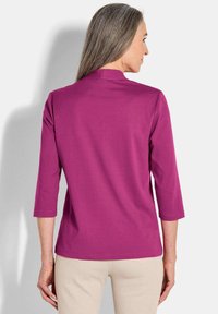 Donna con lunghi capelli grigi che indossa un top magenta a tre quarti e pantaloni beige, in piedi con le spalle rivolte verso di noi su uno sfondo bianco.