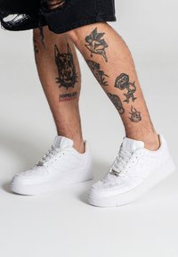 Sneakers bianchi con motivo testurizzato, suola piatta e occhielli argentati. Indossati con pantaloni corti neri strappati; tatuaggi visibili sulle gambe inferiori.