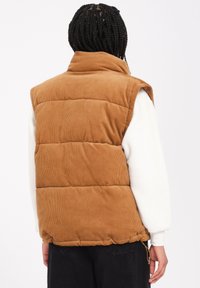 Vesti puffer dari korduroy coklat dengan kerah tinggi, bagian yang dipadati, kantong samping, dan hem yang dapat disesuaikan. Dipakai di atas atasan lengan panjang putih.