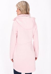 Woman with blonde hair wearing a knee-length, light pink hooded coat viewed from the back against a white background.