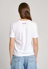 Balta medvilnės marškinėliai su trumpomis rankovėmis, apvaliu kaklu ir raudona "PEPE JEANS" užrašu nugaros pusėje. Sukombinuota su šviesiai mėlynomis aukštu juosmeniu džinsomis.