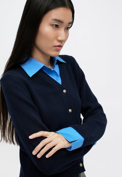 Námořnický pletený cardigan se zlatými knoflíky, nošený přes světle modrou košilovou halenku. Cardigan má design s výstřihem do V a žebrované manžety.