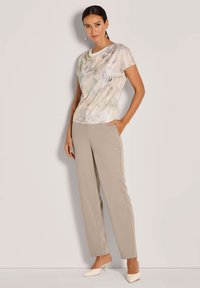 Lichtgekleurde bloemenprint top met geraffineerde halslijn, gecombineerd met beige broek. Het model draagt witte hakken, wat de soepele stoftextuur benadrukt.