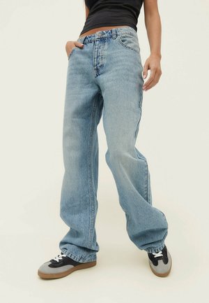 Persona vestida con vaqueros sueltos azul claro, top corto negro y zapatillas negras y grises, de pie con una mano en el bolsillo.