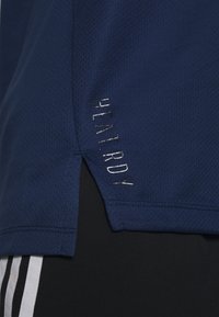 Modrá sportovní košile s prodyšným lemem a textem "HEAT.RDY", nošená přes černé kalhoty s bílými postranními pruhy.