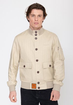 Młody mężczyzna noszący beżową bomberkę zapinaną na guziki, z kieszeniami na klapki i kieszenią na rękawie zapinaną na suwak, w czarnych spodniach.