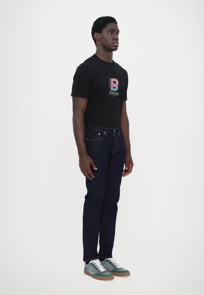 Camiseta de algodón negra con diseño gráfico, jeans de mezclilla azul oscuro y zapatillas de ante verde con cordones blancos y suelas marrones.