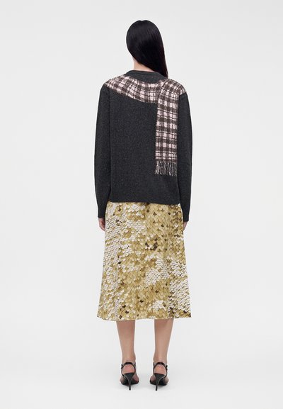 Femme aux longs cheveux foncés porte un pull foncé avec un motif à carreaux roses et une jupe à sequins dorée et blanche, debout en talons sur un fond uni.