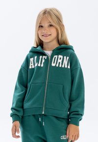Sweat à zip vert avec "CALIFORNIA" imprimé en blanc, présentant une capuche, deux poches avant et des poignets côtelés, associé à un pantalon assorti.