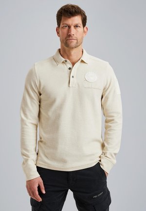 Polo shirt beige à manches longues avec col, patte de boutonnage à trois boutons et logo sur la poitrine gauche ; il présente des poignets côtelés et un ourlet droit.