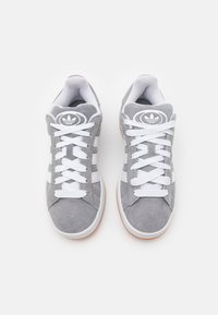 Baskets en daim gris avec des lacets blancs, comportant trois rayures blanches et un logo circulaire sur la languette. Semelle en caoutchouc avec un motif texturé.