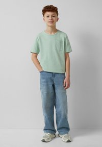 Lichtgroene t-shirt met korte mouwen en een zak, gecombineerd met lichtblauwe denim broeken met een patchwork patroon en neutrale sneakers.